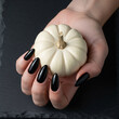 © Vadym - Woman's hand with a perfect glossy black manicure holding a small white pumpkin. Elegant Halloween nail design or autumn beauty salon concept on a dark slate background.