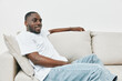 © SHOTPRIME STUDIO - Smiling African American man relaxing on a beige couch in casual white apparel, showcasing comfort and style, ideal for lifestyle and happiness themes
