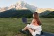 © SHOTPRIME STUDIO - A relaxed woman sits on a blanket with a laptop in a scenic meadow. She wears casual attire and looks focused, enjoying productive remote work in a peaceful outdoor setting.
