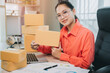 © JK_kyoto - packaging concept. Young woman smiles while showcasing a package in a busy office with stacked parcels and a laptop nearby in a bright environment