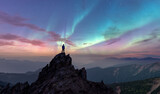 Person Standing On Rocky Peak Under Aurora Borealis Over Mountain Range at Dusk