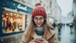 © DaniLana - Young woman wearing a red knitted hat and glasses using smartphone outdoors on a snowy winter day. Warm clothing, snowflakes and city lights create cozy urban atmosphere. Concept of winter lifestyle