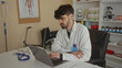 © Krakenimages.com - Young man in a clinic setting working on a laptop, surrounded by medical equipment and supplies, conveying a professional and focused atmosphere.