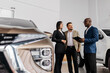 © wedmoments.stock - Group of three individuals, a woman and two men, engaged in conversation at a car dealership, showcasing a modern vehicle and a professional atmosphere of automotive sales