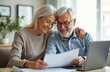 © Lubov - Elderly couple smiles while looking at documents and laptop. They manage finances and plan future investments together at home, showing love and unity.
