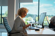 © Videophilia - Senior businesswoman with curly hair sitting at her desk, typing on a computer keyboard and wearing a headset, in a brightly lit modern office environment