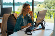 © Videophilia - Young professional woman smiling while working at her desk on a computer, wearing a casual shirt and glasses, in a bright office with green landscape visible outside