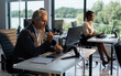 © Videophilia - Mature businessman analyzing data on a computer while working in a modern office, with a female colleague using a keyboard in the background against a bright window view