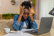 © Andrii Iemelianenko - African American young woman sitting at table on home sofa nervously checks utility bills using laptop, printed papers. Black girl looks stressed overwhelmed trying to manage household payments online