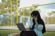 © Fajar - A young woman in a hijab uses her phone and laptop outdoors in a park, appearing focused on her work or studies amidst a natural setting
