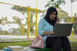 © Fajar - A young woman in a hijab uses her phone and laptop outdoors in a park, appearing focused on her work or studies amidst a natural setting