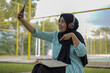 © Fajar - Young woman in hijab and headphones using a smartphone to record a video outdoors with a notebook and pen nearby, capturing a moment of content creation and learning in a casual setting