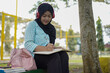 © Fajar - Serene young woman in a hijab and red headphones, deeply engrossed in reading a book while studying outdoors on a tranquil park bench, fostering knowledge and personal growth