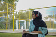 © Fajar - Serene young woman in a hijab and red headphones, deeply engrossed in reading a book while studying outdoors on a tranquil park bench, fostering knowledge and personal growth