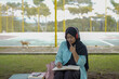 © Fajar - Serene young woman in a hijab and red headphones, deeply engrossed in reading a book while studying outdoors on a tranquil park bench, fostering knowledge and personal growth