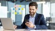 © StocksJust4You - Smiling man multitasking at his desk, using smartphone while working on laptop in modern office with professional atmosphere