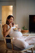 © Drazen - Happy pregnant woman eating cookies while relaxing in armchair at home.