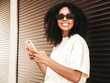 © halayalex - Beautiful woman with afro curls hairstyle.Smiling hipster model in white t-shirt. Sexy carefree female posing in the street near brown wall in sunglasses.Looking at smartphone screen, using apps