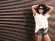 © halayalex - Beautiful woman with afro curls hairstyle.Smiling hipster model in white t-shirt. Sexy carefree female posing in the street near brown wall. Cheerful and happy, in sunglasses