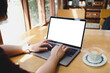 © Thaspol - A person working on a laptop with a blank screen at a sunlit wooden table in a cafe, enjoying a cup of coffee. A concept for remote work or freelancing.