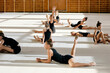 © pressmaster - Group of Caucasian teenage girls stretching on gym floor during rhythmic gymnastics training, performing various flexibility exercises with focused expressions in spacious sports hall