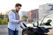 © sofiko14 - A man in glasses uses his phone while charging his electric car at a charging station in an urban setting.