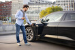 © sofiko14 - A man connects a charging cable to his black electric vehicle in an urban setting, holding his phone.
