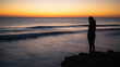 © faisal - Figure standing near ocean cliff watching sunset, waves softly blurred with long exposure, warm gradient sky meeting horizon, faceless silhouette composition, cinematic travel mood