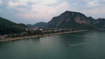 Wall Mural - Aerial view of Ao Nang Beach at dusk with calm sea, limestone cliffs, longtail boats, and lively town lights beginning to glow.