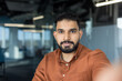 © Liubomir - Young indian man in brown shirt confidently taking a selfie in a modern office, creating professional social media content and showcasing entrepreneur style and smart-casual confidence