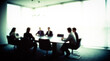 © Dmytriy - A blurry, grainy image of a group of people sitting around a table in a bright conference room, giving the impression of a business meeting or conference