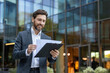 © Liubomir - Confident businessman reviewing important papers on a clipboard with a content smile, standing in front of a modern glass office building, symbolizing success and corporate life