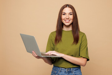Young smiling happy IT woman she wears olive green t-shirt casual clothes hold use work on laptop pc computer isolated on plain pastel light beige color background studio portrait. Lifestyle concept.