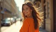 © miss irine - Young woman in orange top walks on city street looking back and smiling. Her hair blows in wind. Sunlight brightens scene. She looks happy and playful outdoors.