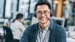 © mila103 - A friendly man wearing glasses and a headset smiles while assisting clients from his well-organized office. Nearby another employee is also engaged in their work creating a collaborative atmosphere.