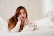 © Thitiporn - Tired Asian woman lying on bed with sad face holding alarm clock. Concept of sleep problem, insomnia, stress, or difficulty waking up. Morning routine struggle in cozy bedroom setting.