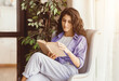 © Prostock-studio - Young woman sits comfortably in an armchair, engrossed in a book. Sunlight streams through the window, creating a warm atmosphere in her stylish living room filled with green plants.