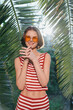 © SHOTPRIME STUDIO - A cheerful young woman stands outdoors among tropical plants, wearing a red striped crop top and orange sunglasses. She smiles warmly, radiating sunshine, confidence, and carefree summer energy in a