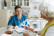 © shurkin_son - Smiling caring healthcare worker in blue uniform answering questions of her aged female patient, retired woman showing her blister with pills while sitting in kitchen. Consulting with doctor at home