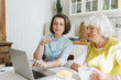 © shurkin_son - Indoor image of social worker and her senior female patient sitting at kitchen table, caregiver teaching retired person how to use laptop and wireless internet and applications remotely online