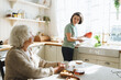© shurkin_son - Caring mature female caregiver helping her senior patient to cook, standing next to counter holding pumpkin, going to bake pie while talking to elderly woman sitting at kitchen table