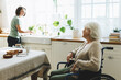 © shurkin_son - Senior lady in wheelchair in kitchen talking to nurse cooking dinner or washing dishes. Social worker helping retired disabled female with domestic chores at home, having warm conversation