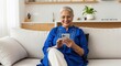 © PRASANNAPIX - Happy senior Indian woman using smartphone on sofa at home.