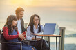 © shine - Small diverse business team of three colleagues meeting at laptop, watching online startup presentation, website content, talking on rooftop near the sea. Colours of sunset.Skyscape nature composition