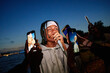 © Seventyfour - Young Black man singing into microphone outdoors at dusk surrounded by multiple hands holding smartphones recording and taking photos, waterfront and city lights in background