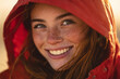 © EricMiguel - Close-up portrait of a freckled young woman smiling warmly beneath a red hooded jacket, golden sunset backlight, natural makeup and wind-swept hair capturing joyful outdoor mood