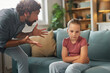 © Stockphotodirectors - Father is visibly upset as he gestures animatedly, while his daughter sits with crossed arms, appearing withdrawn and contemplative in their comfortable living space.