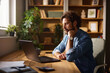 © Stockphotodirectors - A man is seated at his wooden desk in a home office. He is intently looking at his laptop while resting his chin on his hand. Shelves filled with books and plants surround him.