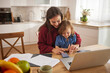 © Stockphotodirectors - A mother enjoys quality time with her child while helping with a project at the kitchen table, surrounded by papers, fruits, and a laptop. The atmosphere is warm and loving.