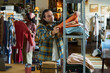 © pressmaster - Middle aged Caucasian man organizing folded clothes on metal shelf while Black woman browsing hanging garments in thrift shop, both focused on sorting and selecting secondhand clothing items